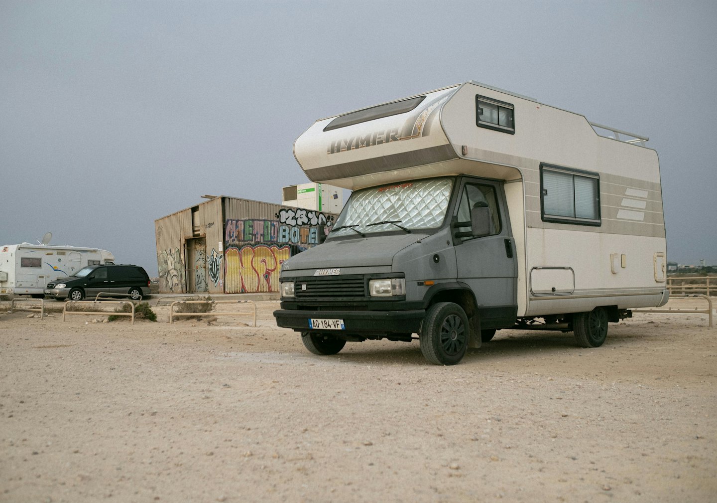 Grey and white campervan parked on gravel lot with graffiti building.