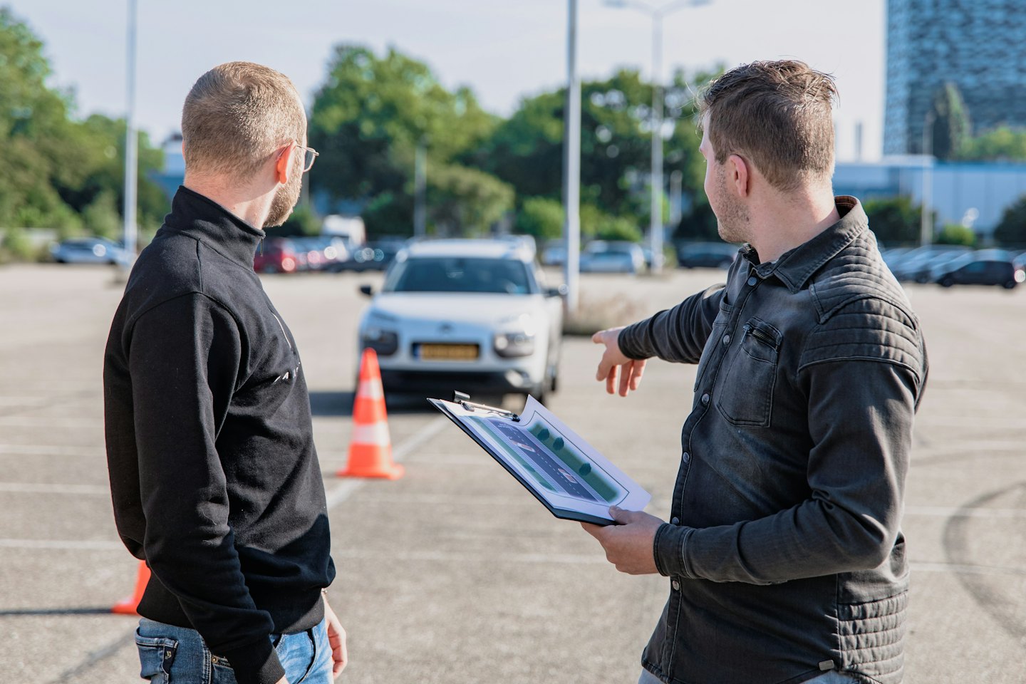 Driving instructor explains diagram on clipboard next to car and cones.